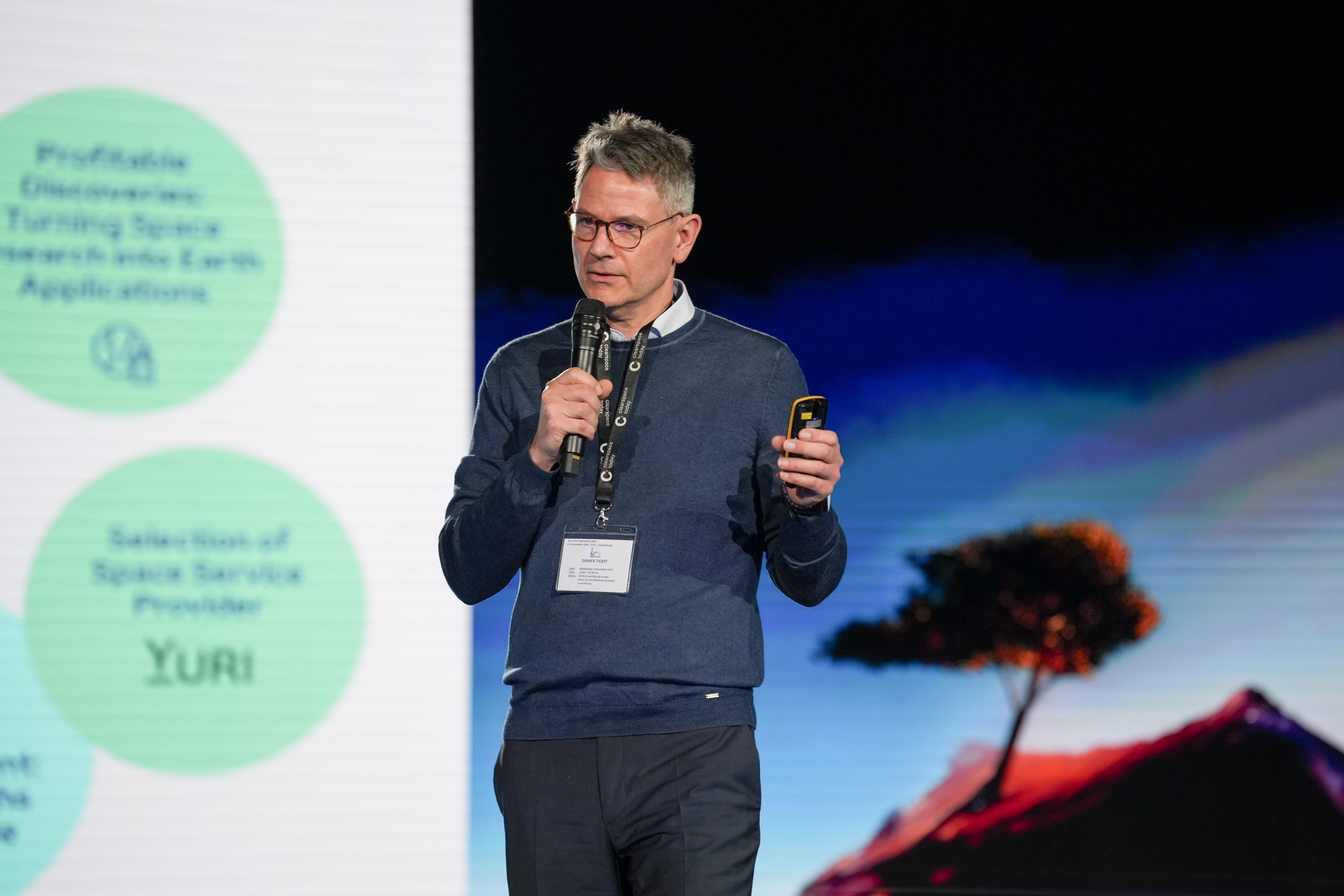 A white man in his early fifties wearing glasses, a blue sweater and blue pants on stage holding a microphone, giving a presentation.